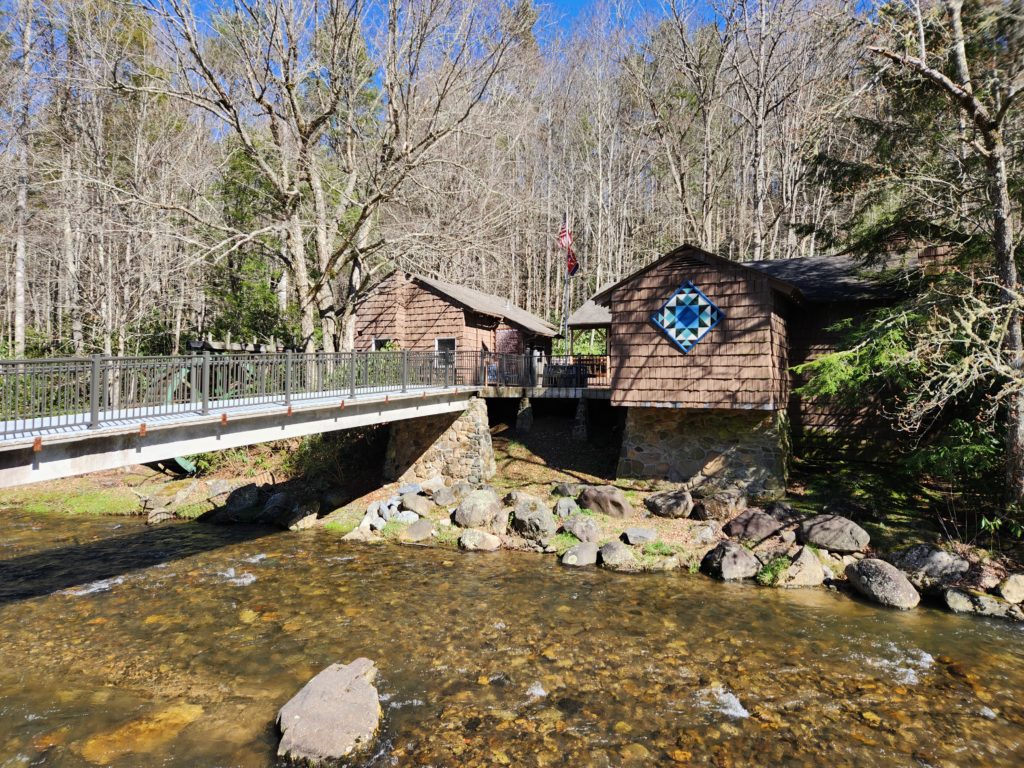 A brown building with wooden siding and a metal bridge that goes over a river leading to it. The visitor's center at Roan Mountain State Park in TN.