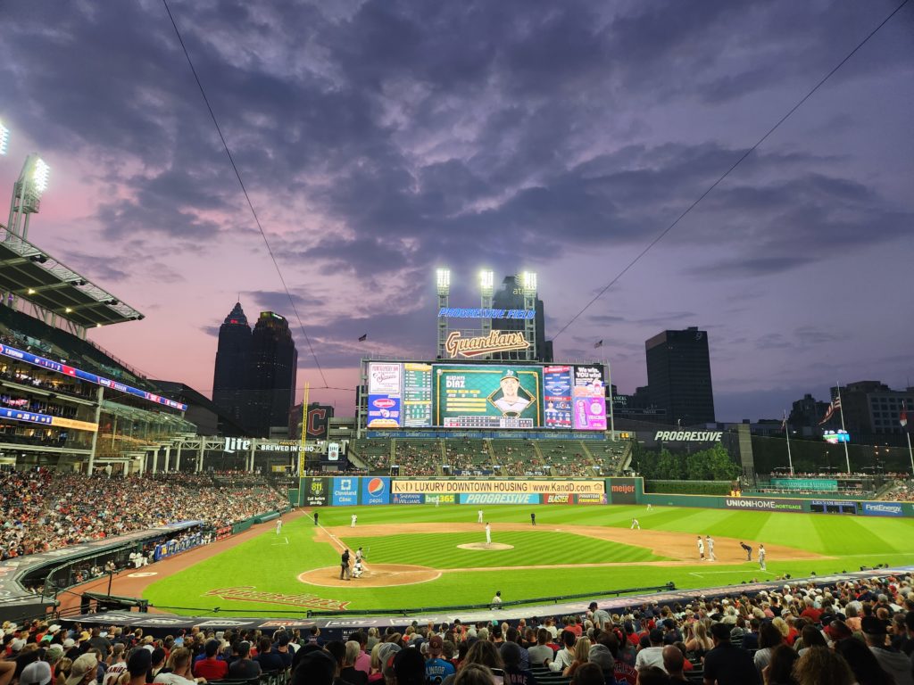 The Cleveland Guardians Progressive Stadium. A game is being played and the setting sun makes the sky in the background look purple, blue, and pink. 