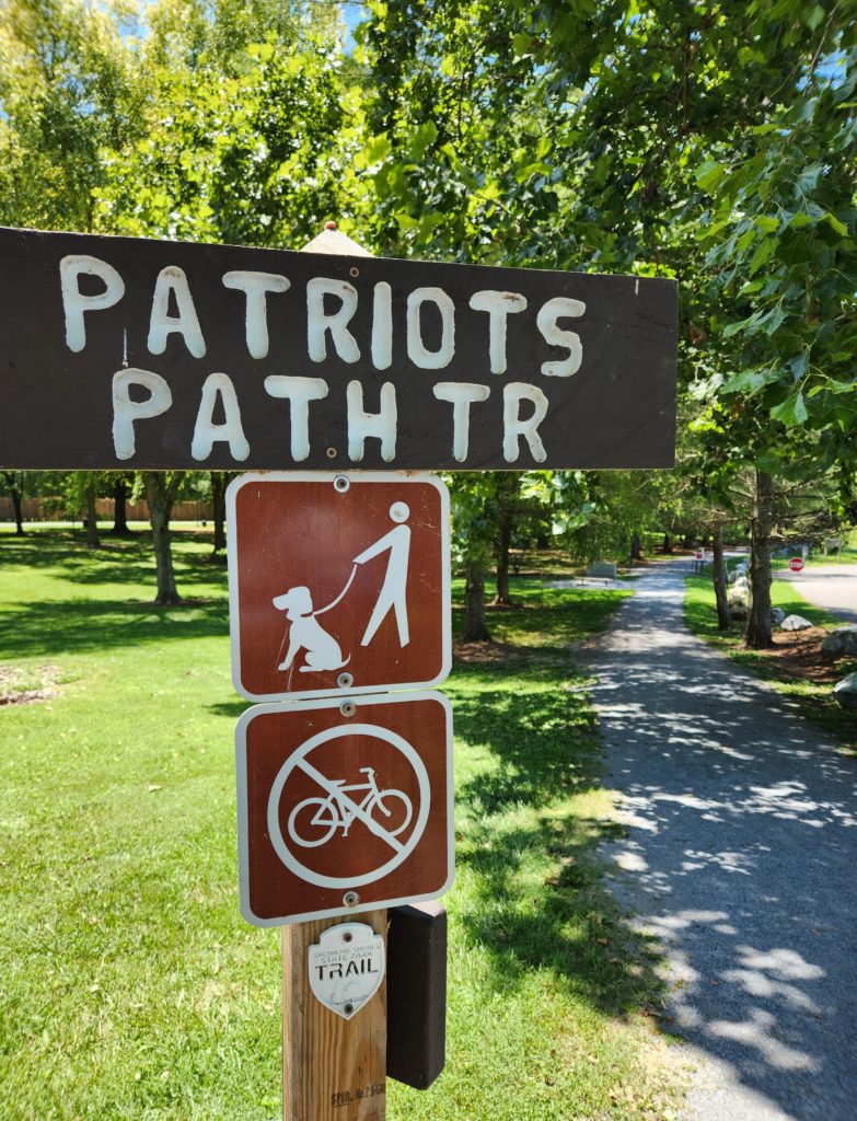 A sign that reads "Patriots Path Tr" (trail), one that reminds people to keep their dog on a leash, a no biking sign, and a small trail marker that reads "Sycamore Shoals State Park Trail", all on a wooden post at the beginning of a gravel covered trail. 