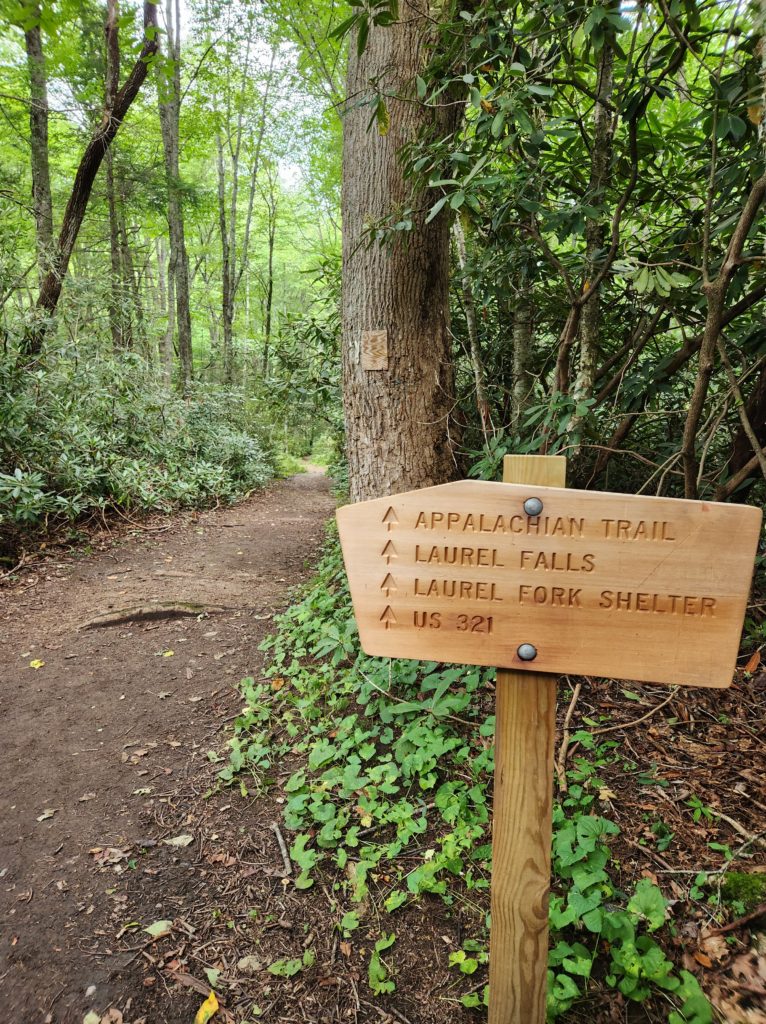 A dirt path leading into the forest, with a wooden sign on the right side that points the way to the Appalachian Trail, Laurel Falls, Laurel Fork Shelter, and U.S. 321 in TN.