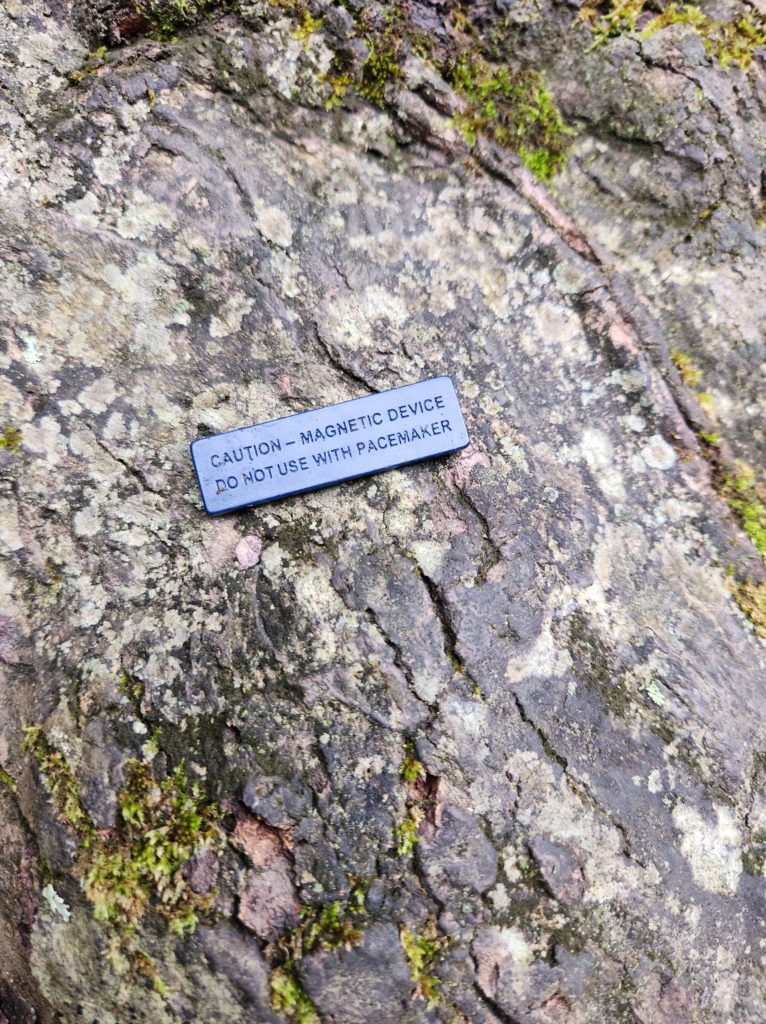 A small, dark blue magnet laying on a rock. The magnet reads, "Caution - magnetic device do not use with pacemaker". 