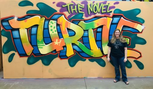 A wall with colorful graffiti on it that says "The Novel Turtle" with the creator standing in front of it.