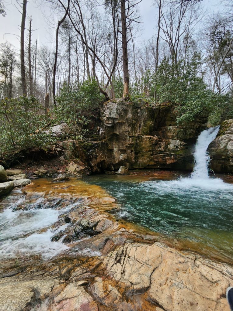 Blue Hole Falls in Elizabethton, TN. Aptly named because the water flows over the rocks into a "hole" below. The water is blue-green in color. Brown rocks, trees and bushes are all around it. A blue sky shows through the trees in the background.