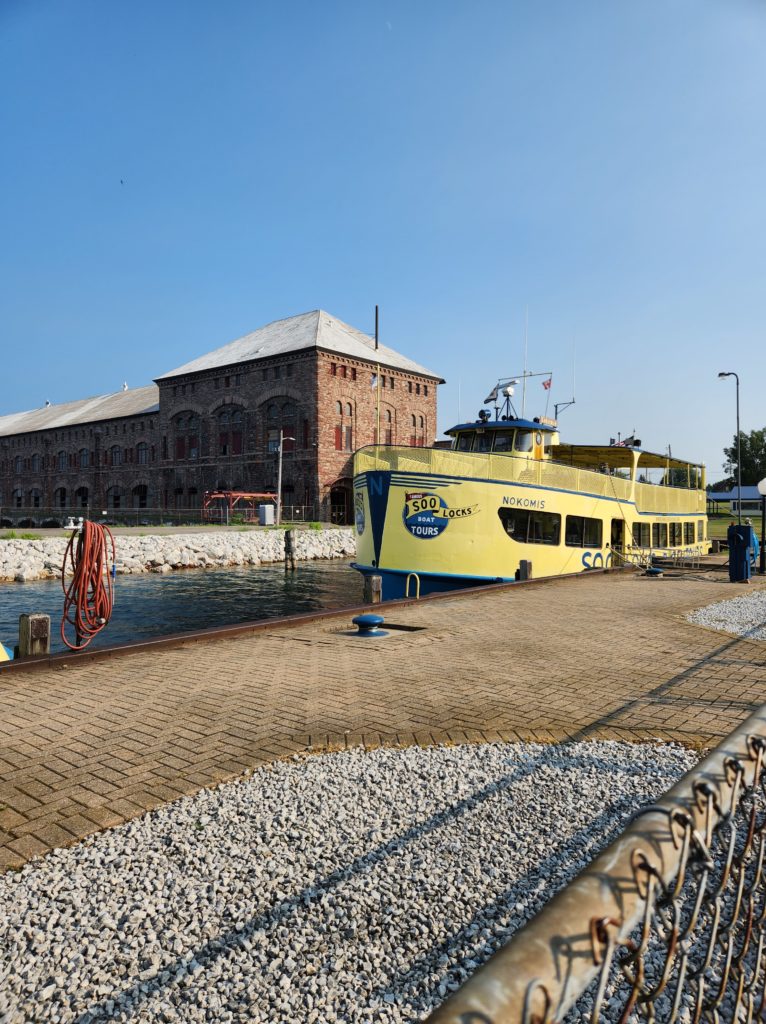 The "Famous Soo Locks Boat Tours" yellow boat sitting at a concrete dock. A large brick building in the background. 