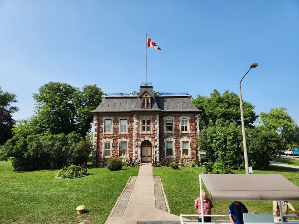 A stone building with the Canadian flag flying on top. It's surrounded by green grass, trees, and bushes. A sidewalk leads down to a covered booth where the Soo Locks operator is.