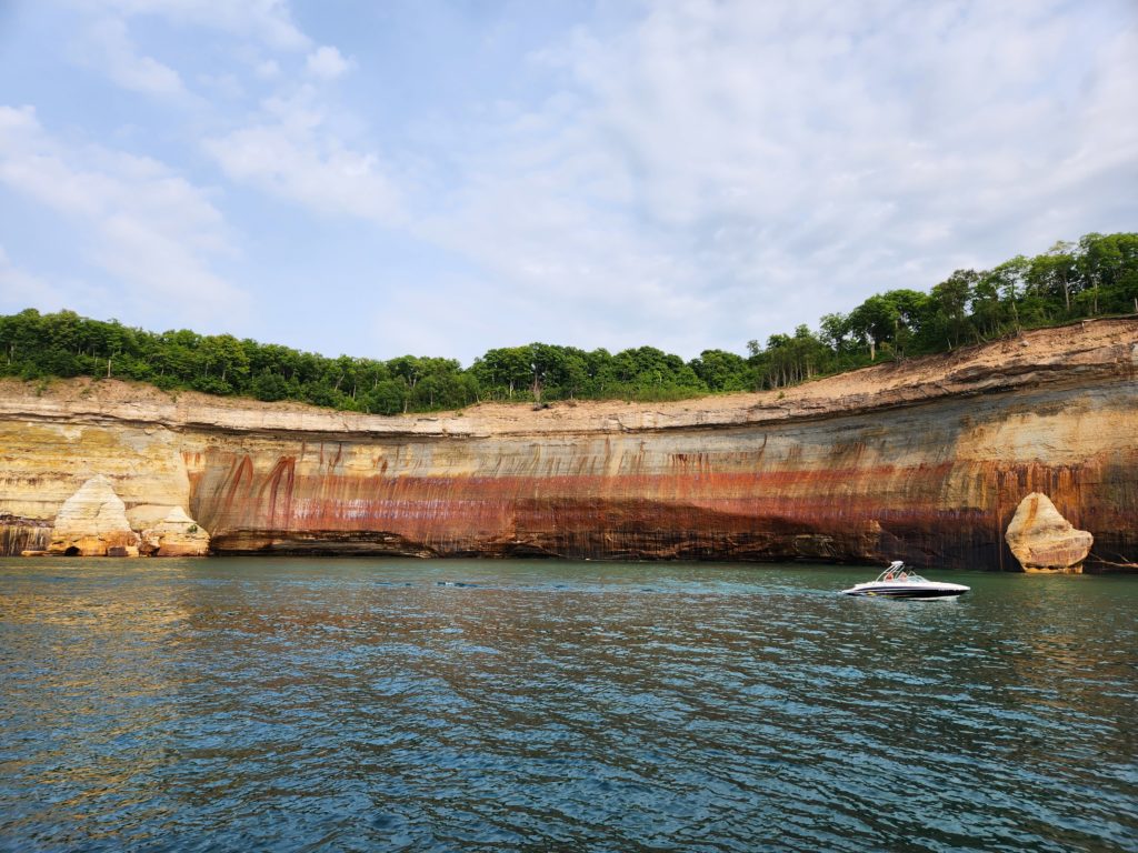 A boat going by the rock wall that is Pictured Rocks National Lakeshore. The boat is in Lake Superior, and there is blue sky with a few clouds at the top of the picture. 