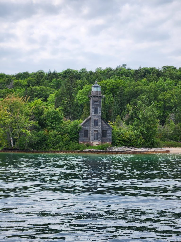 The Grand Island East Channel Lighthouse in Michigan. Blue sky and a few clouds line the top of the picture. A small dark colored wooden lighthouse sits among all different types of trees in the center. Along the bottom of the picture is Lake Superior. 
