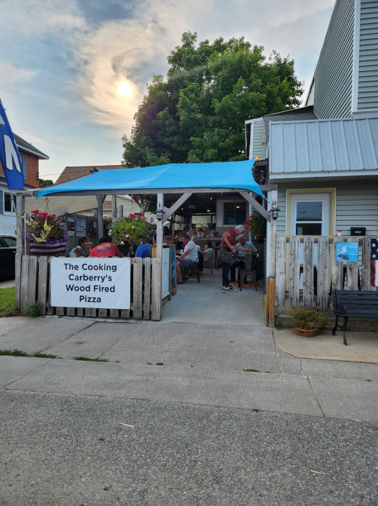 A picture of an outdoor space with a pergola type roof with a blue covering. People are sitting in that space. The sign reads "The Cooking Carberry's Wood Fired Pizza". From the perspective of someone standing across the street. 