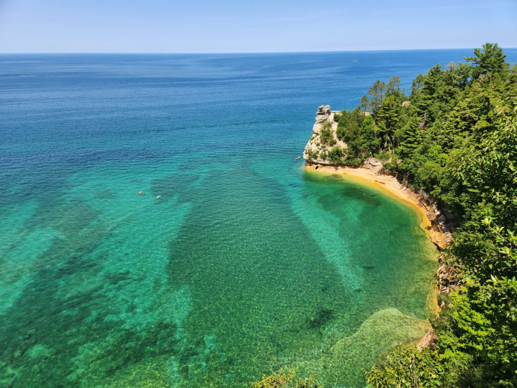Looking down from a high viewpoint to the waters of Lake Superior below. Part of the Pictured Rocks National Lakeshore shoreline runs along the right side of the water, with beach at the bottom and trees all over the hill. 