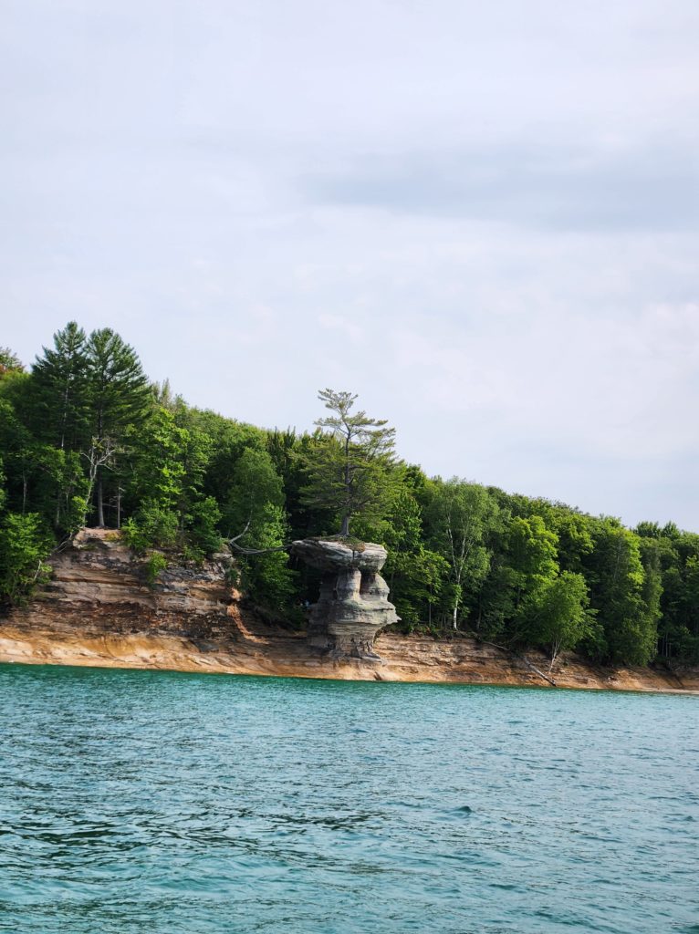 Chapel Rock at Pictured Rocks National Lakeshore. A famous rock formation that juts out from the trees and rock wall. On the rock, there is a tree growing, with its roots stretching back towards the land across the way. Lake Superior lines the bottom of the picture. 
