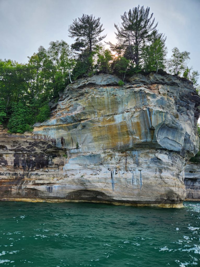 One of the rock formations at the Pictured Rocks National Lakeshore. Blue sky is at the top of the picture, green trees with the sun peeking through line the top of the rock. Then below the rock, Lake Superior. 