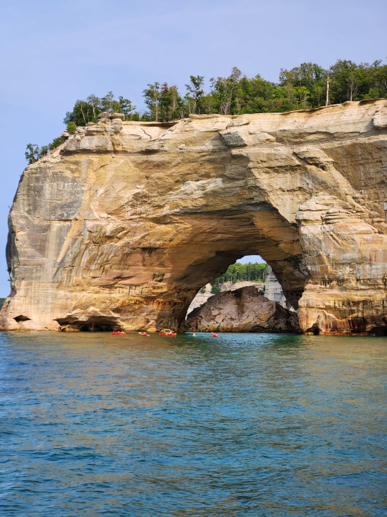 The famous arched rocked formation at Pictured Rocks National Lakeshore. Blue sky and a few trees line the top of the picture, and Lake Superior lines the bottom. 