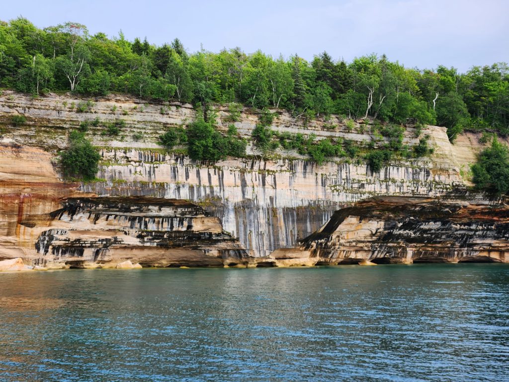 A section of the Pictured Rocks National Lakeshore in Michigan. Blue sky, then green trees, the gorgeous rock formation with black and white colors dripping down the side of it like paint dripping off of a canvas, and Lake Superior at the bottom. 