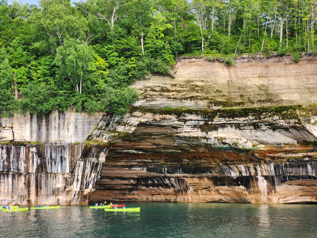 A rock formation at the Pictured Rocks National Lakeshore that creates a cave. Blue sky and trees are at the top of the picture. The rock wall and cave are across the middle, with black and white "painted" rocks. Lake Superior lines the bottom, with a few kayakers in brightly colored kayaks floating in the water. 