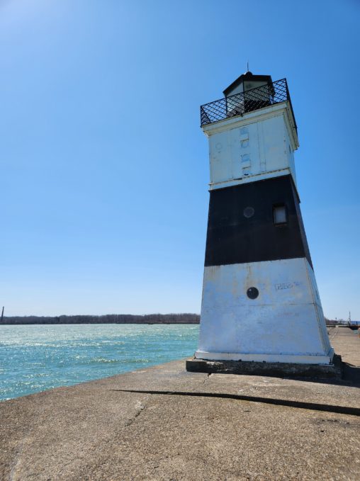 A small, square, metal, black and white lighthouse sitting at the end of a wide concrete dock. Lake Erie is in the background.