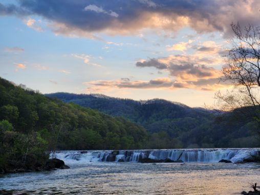 Sandstone Falls in WV. A wide waterfall runs through the middle of the picture. It is surrounded by the mountains and trees. The setting sun is above the mountains in the distance.
