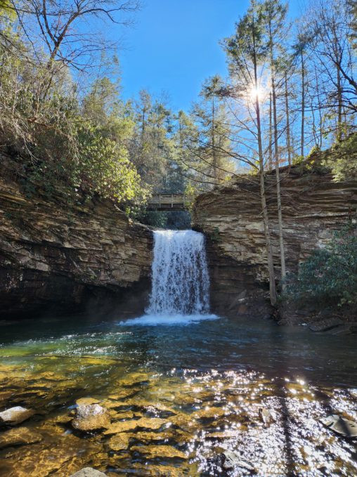 Little Stony Falls in Dungannon, VA. A waterfall flowing over a layered rock wall, into a river below. Trees all around. The sun is peeking through the trees.