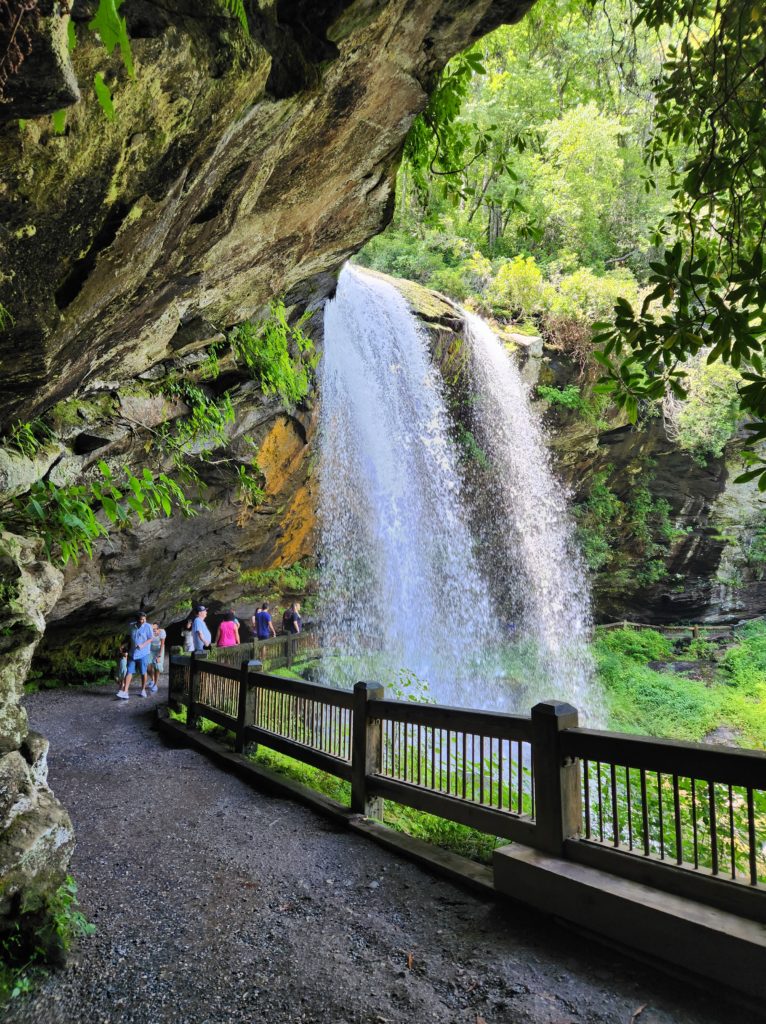 Walking behind Dry Falls in NC. A gravel pathway with a small wooden fence, rocks on your left and the back of the waterfall on your right. 