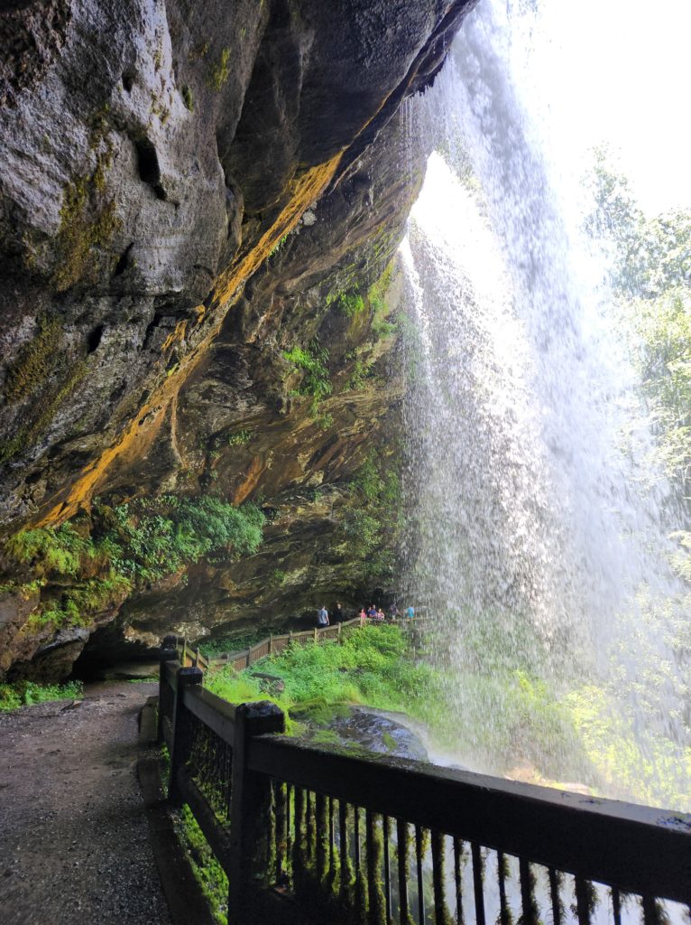 Walking behind Dry Falls in NC. A gravel pathway with a small wooden fence, rocks on your left and the back of the waterfall on your right. 