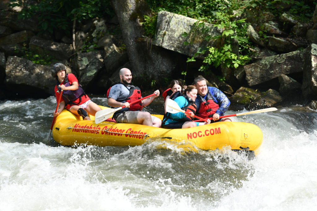 Five people white water rafting. Three of them are leaning inwards, towards the center of the boat. one of them is sitting upright. And the guide in the back of the boat is leaning hard to the left to direct the raft. 