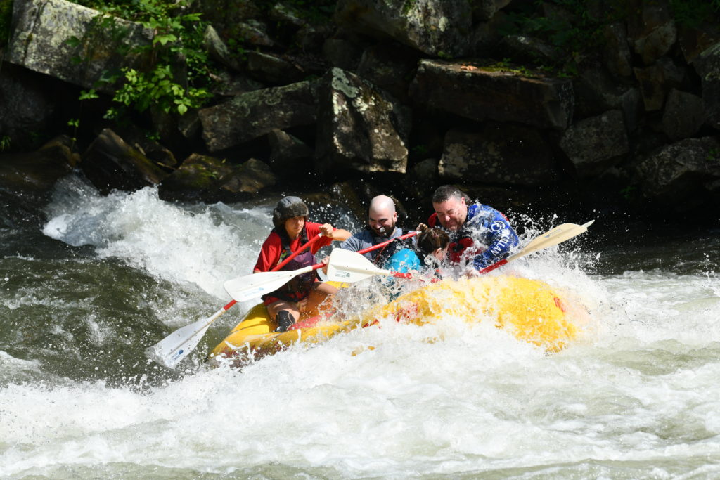 Five people who are white water rafting, going over a rapid. It looks a little more like they are going through AND over it because they are surrounded by the splash of water. 
