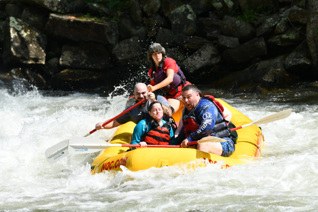 Five people in a white water raft, coming out the other side of a rapid. The front two are drenched, one is hidden behind the person in front of them, and the other two (including the guide) are directing the boat with their paddles. 