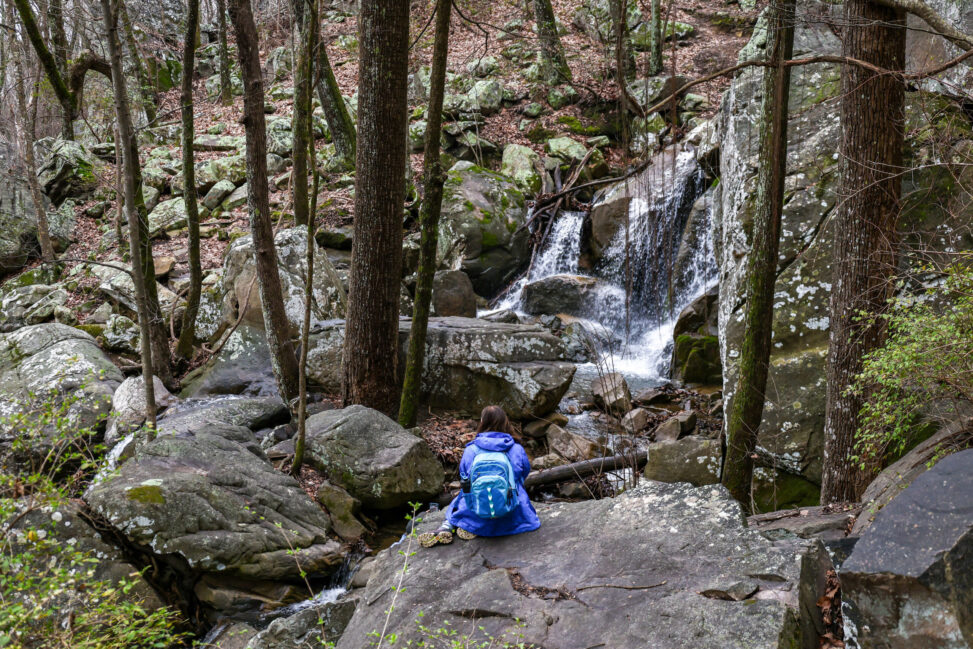 A woman sitting on a large boulder looking at a waterfall. Her back is to the camera, she's wearing a jacket and a backpack, and is surrounded by trees and large rocks.